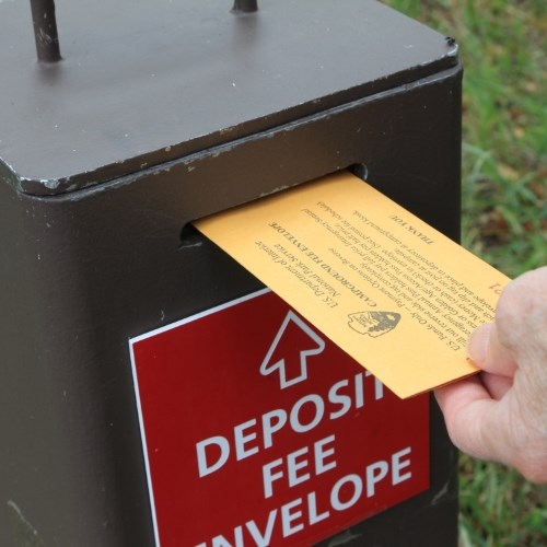 A hand puts a yellow envelope into a metal box. The box has a red "Deposit Fee Envelope" sign.