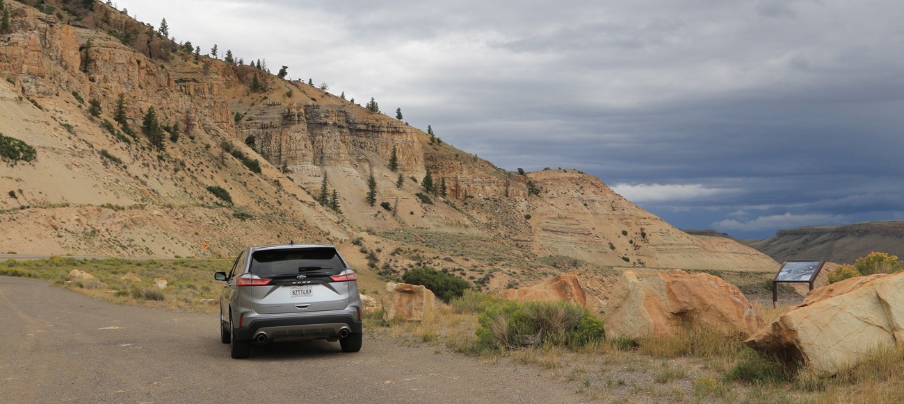 A grey SUV parked on a gravel pulloff. A wayside is off the side of the road; geologic formations in the background.