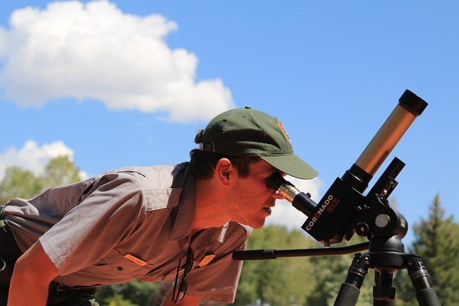 A park ranger wearing a grey shirt and green ball cap looks through a small mounted telescope.