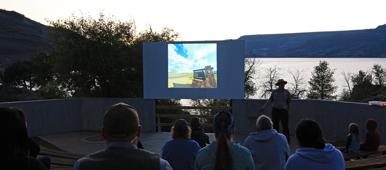 A park ranger stands in front of a seated group at an amphitheater. People are looking at a screen with an image. A reservoir and mesas are visible on the horizon.