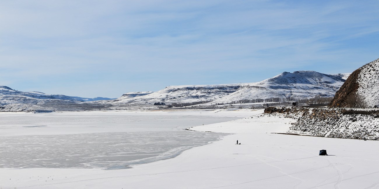 Landscape with snow, frozen reservoir, and mesas surrounding it. A person and tent are silhouetted on the frozen surface.