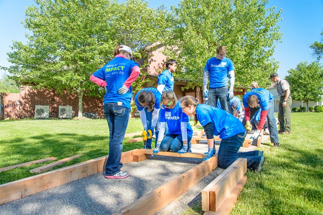 A group of 10 people in blue shirts stand and kneel around wooden frames; two use yellow drills as the others hold the wood in place.