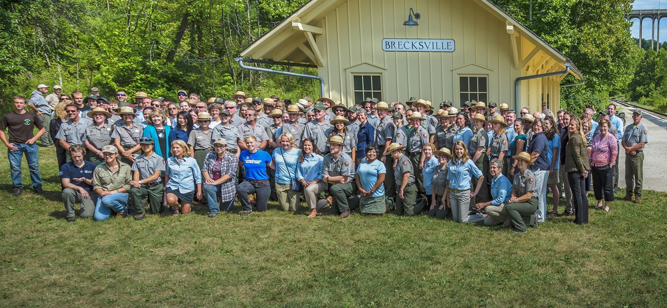 Group photo of 130 people standing and kneeling in rows in front of a yellow building with a sign that reads “Brecksville”. Most wear the green and gray National Park Service uniform; several others wear light blue shirts and khaki pants or skirts.