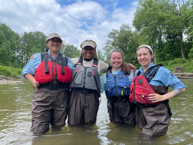 Four young people in waders and life vests stand smiling in knee-deep water.