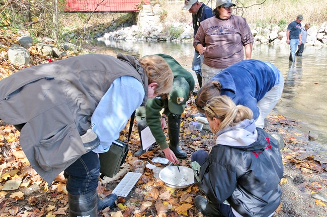 Uniformed ranger and 3 others gatherd around a container with water with 3 others are wading in a stream in the background