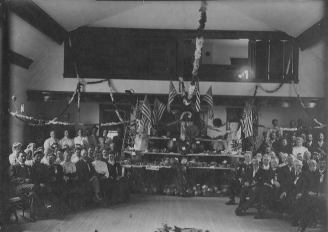 Black and white photo of about 75 people seated inside a room with a large display of flags and pumpkins on tables in the middle.