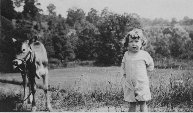Black and white photo of a young girl standing next to a cow in a farm field.
