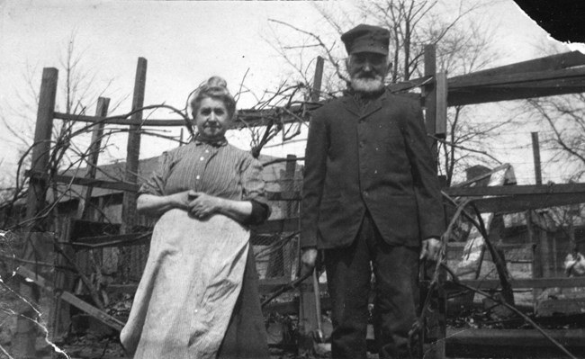 Black and white photo of a woman wearing an apron and a man in buttoned coat and hat, standing in front of a wooden fence.