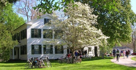 Exterior of a two-story white house with green shutters; a tree with white blossoms stands in front of and as tall as the house; a dozen people and bicycles stand in the grass and ride along the driveway next to the house.