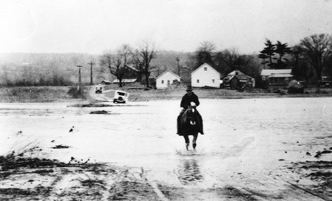 Black and white photo of a man on horseback riding through floodwaters down a flooded street.