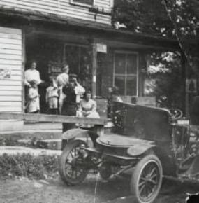 Historic photo of a family of six stand on the porch of their home with an old model automobile sitting in the foreground.