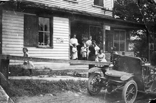 Black and white photo of a family of eight standing on the porch of their home with an old model automobile sitting in the foreground.