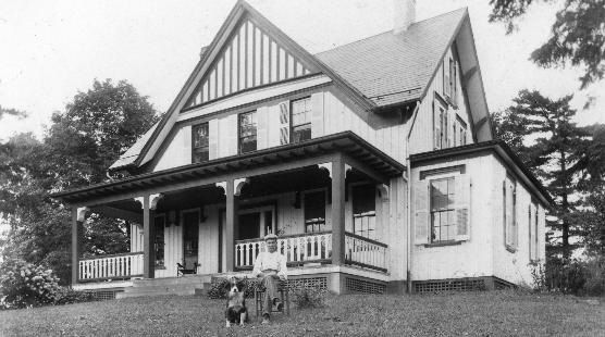 Black and white photo of a two-story house with pointed roof and striped siding; a man sits in a chair next to a dog in the foreground.