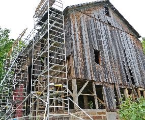 Scaffolding is stacked up along the side of a brown wooden barn while the framing is being replaced.