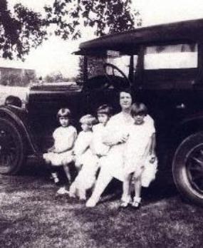 Historical photo of woman and four children sitting on the running board of an 1920's style automobile.