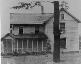 Black and white photo of Nathanial Point farmhouse, a two-story home with an enclosed porch and a large tree in the front yard.