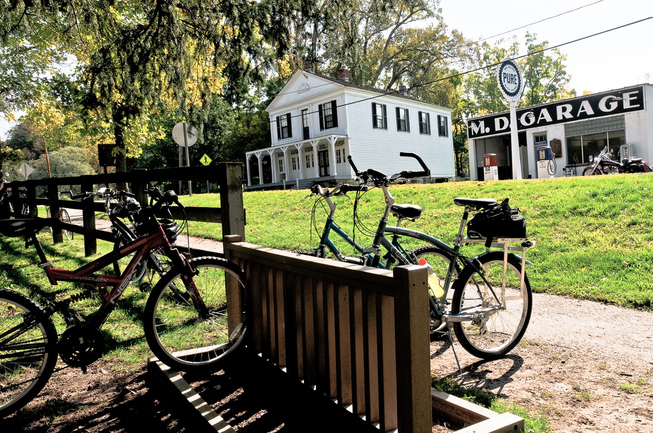White, two-story building with black shutters and covered porch; next door, a white single-story building with large letters, "M.D. Garage" painted in black and white; in the foreground three bikes at a bike rack near the end of a wooden fence.