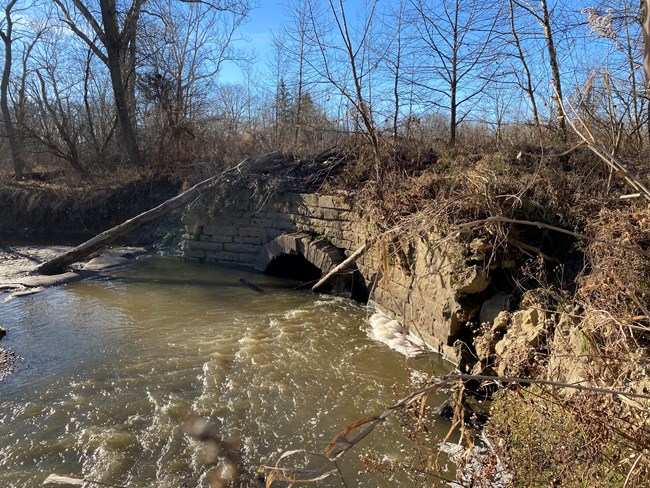 A gray stone wall with small arched opening near the bottom, half submerged in brown water; trees and shrubs grow around it.