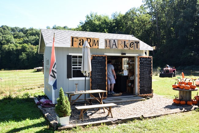 A small shed-like structure in front of a green fenced field, with sign along the roof line, "Farm Market"; a person stands inside and the double doors are open with black chalkboards on each listing products and prices.