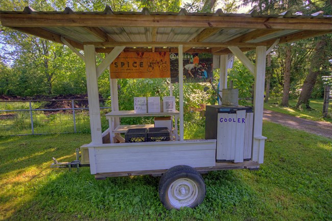 A wooden farm stand with a tin roof sits on a trailer; a sign in the upper left corner reads, "Spice Acres" and another on a mini-fridge near the bottom right reads, "Cooler."