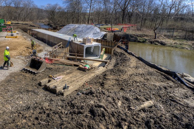 A large, gray, rectangular concrete culvert creates a tunnel beside a creek; three workers in safety gear do various tasks around it.