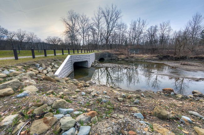 A stream passes through two side-by-side culverts under a trail; one is new and rectangular, the other older and round.