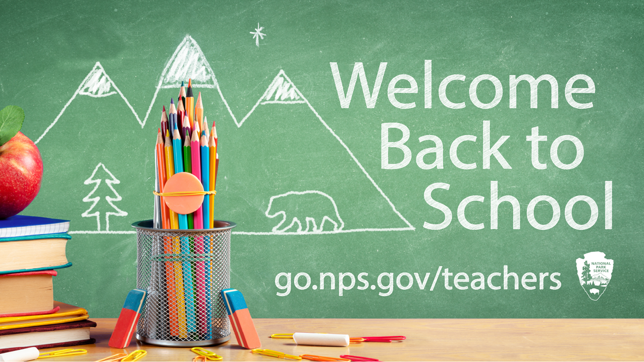 A green chalkboard behind a classroom desk topped with stacked books, an apple, and colorful art supplies. On the chalkboard is a drawing of mountains, a tree, and a bear and the words “Welcome Back to School / go.nps.gov/teachers” and the NPS arrowhead.
