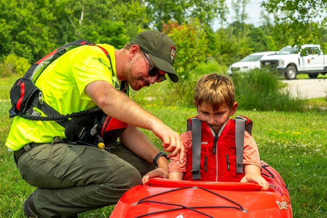 Uniformed river ranger leaning over a kayak with child in it on a grassy area.