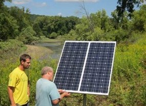 Two people stand next to solar panels next to a stream.