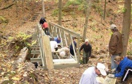 A group of people work along the length of a partially complete fiberglass bridge over a stream in a forest.