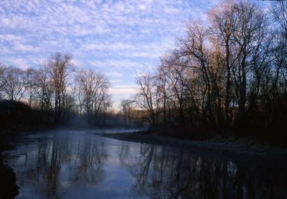 A river at dusk reflects silhouettes of leafless trees and a blue sky.