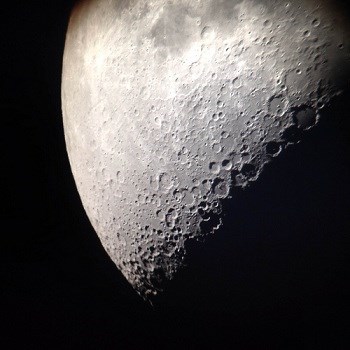 A zoomed in photo of a half moon glowing white with its cratered surface in view.