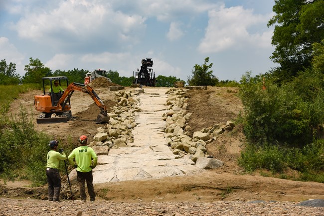 An earth and stone ramp under construction; workers in bright green shirts use heavy machinery to move soil.