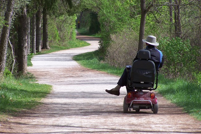 A person in a wide-brimmed hat rides a motorized scooter along a wide stone path through trees.