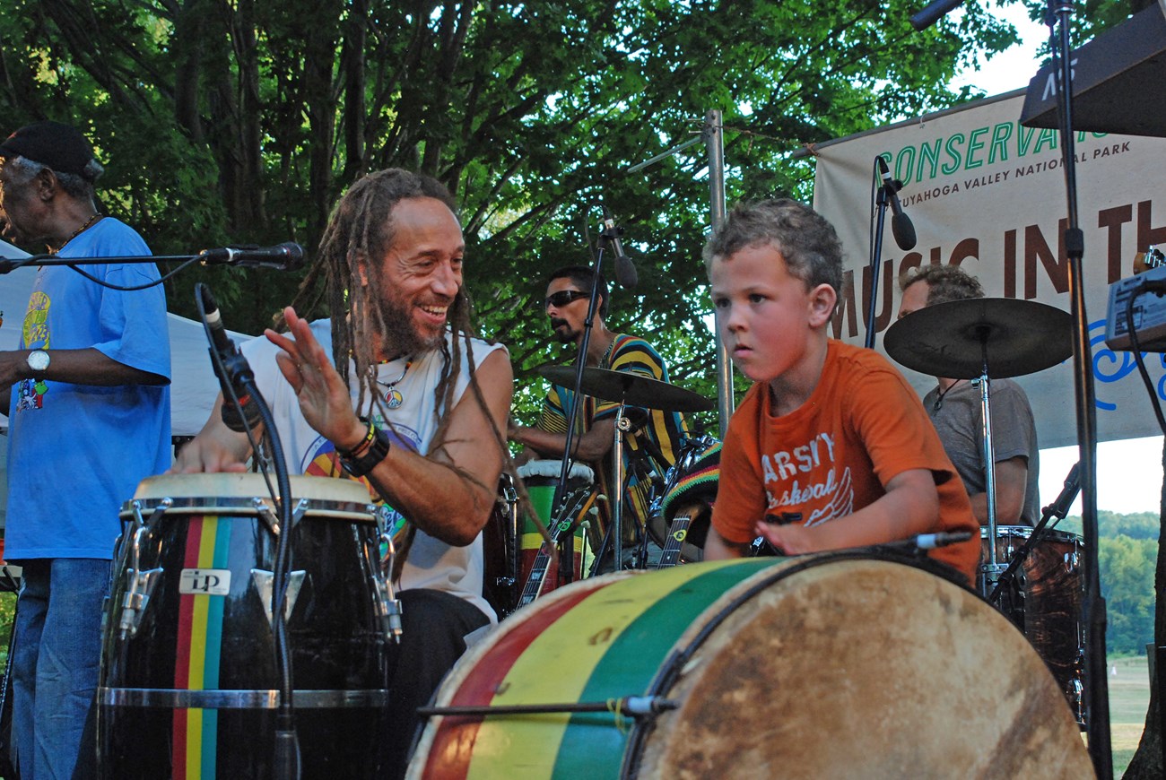 At the front of an outdoor stage under a tree, a musician with dreadlocks drums with his hands and smiles at a child who concentrates on beating a very large drum.