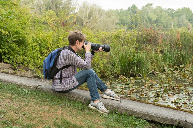 A woman sits on the grass, with her camera ready. A marshy area is in front of her.