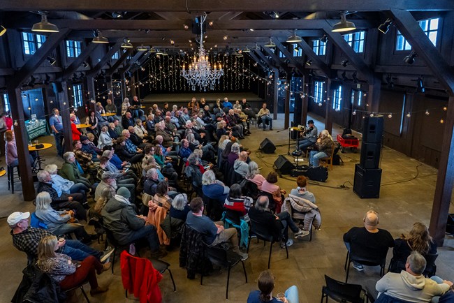 About a hundred people sit in a semi-circle in a large room while two musicians play.