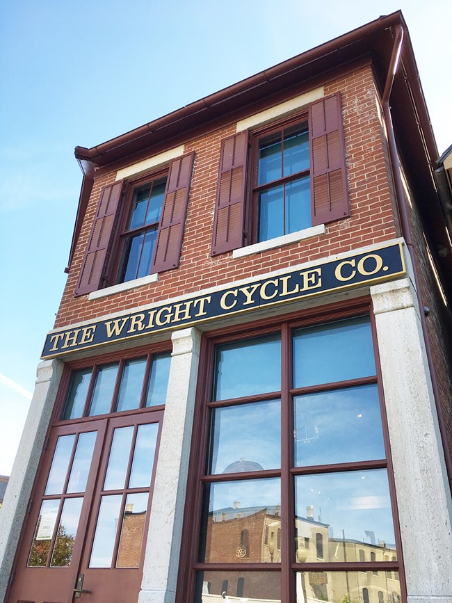 Upward view of the Wright Cycle Company fourth shop. The red brick building has shutters on the second floor windows, and large first floor windows. The reflection of buildings are visible in the windows.