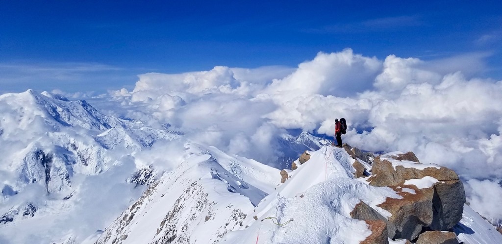 A roped climber stands on a steep rocky ridge above the clouds