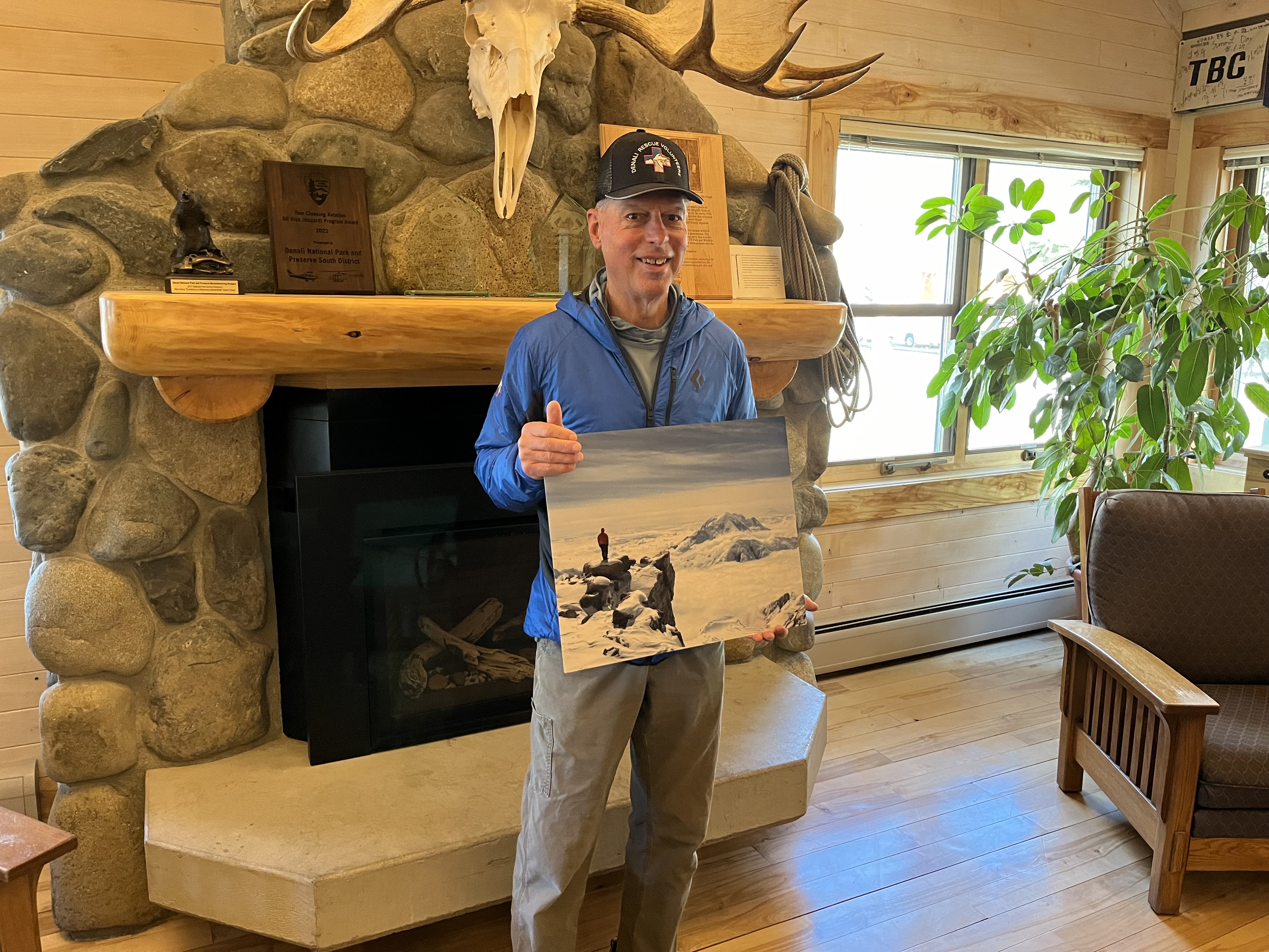 A smiling man in a baseball cap holds a photographic print in front of a stone fireplace