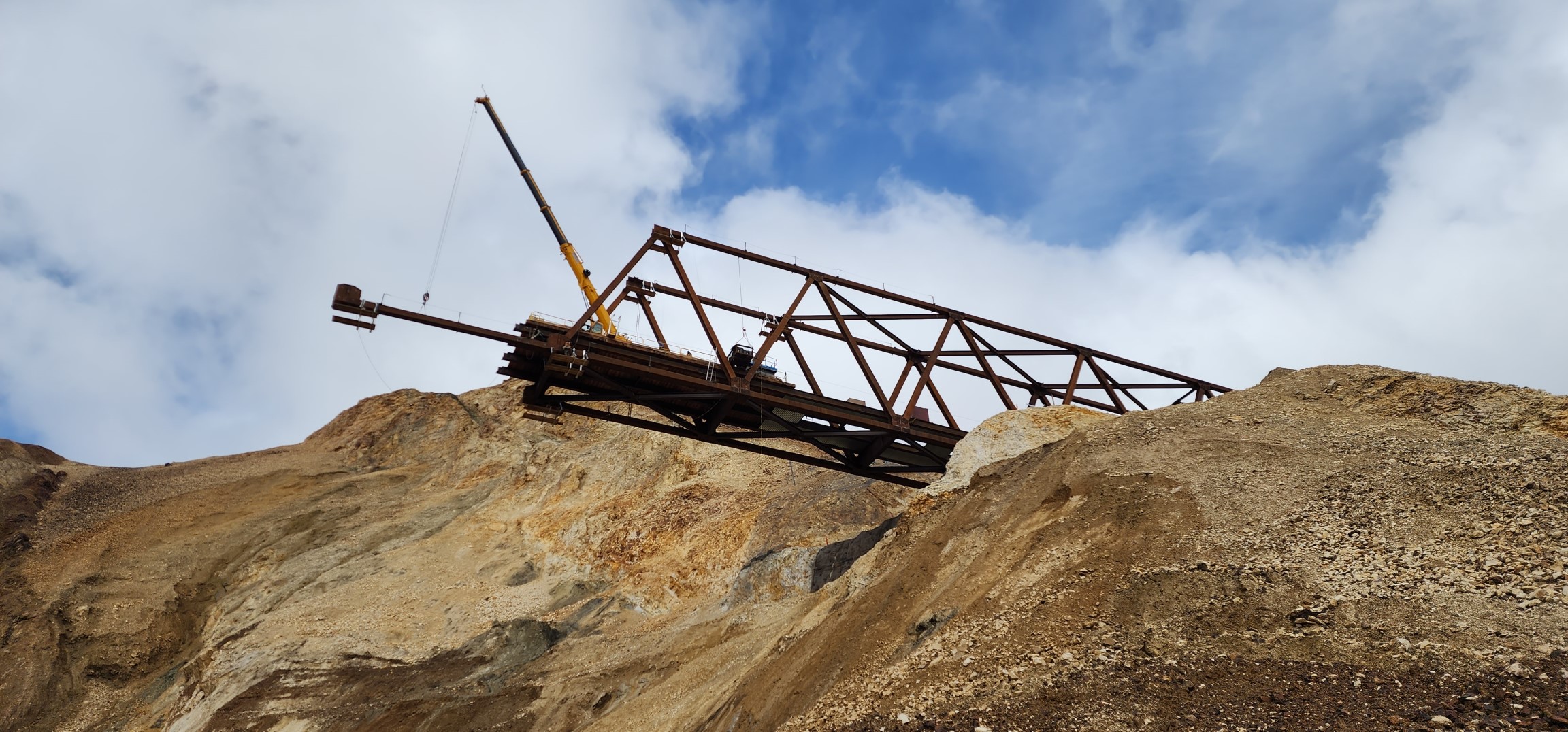 The partial bridge that has been constructed extends out over the landslide and a steep, rocky mountainside. A crane places a metal beam at the end of the bridge.