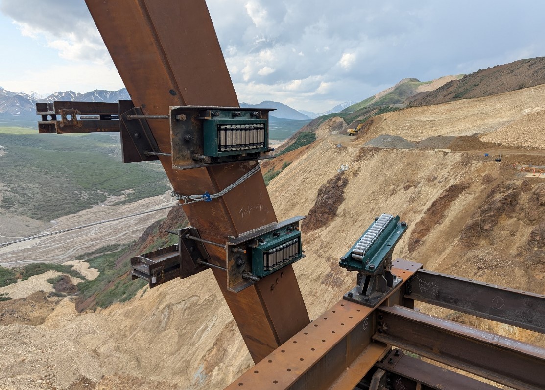 Three rollers attached to metal beams. The mountainside and river valley can be seen far below the metal beams.