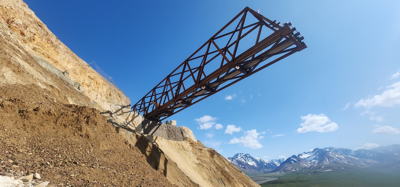 Several trusses of the bridge extend out over the landslide above the viewer against a bright blue sky.