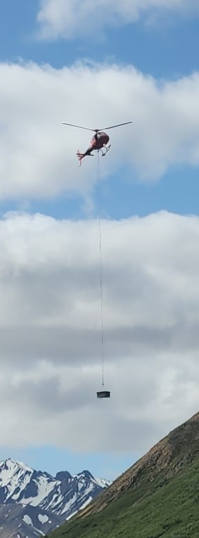 A helicopter carries a large box attached to a long line below the helicopter.