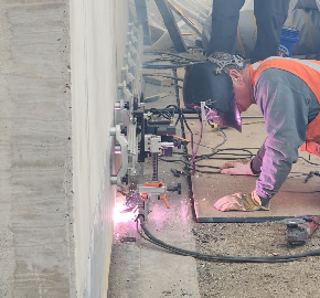 Sparks fly as a welder wearing a full face helmet kneels next to the back wall of the bridge abutment.