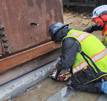 Two works wearing safety vests and helmets kneel next to large metal beams.