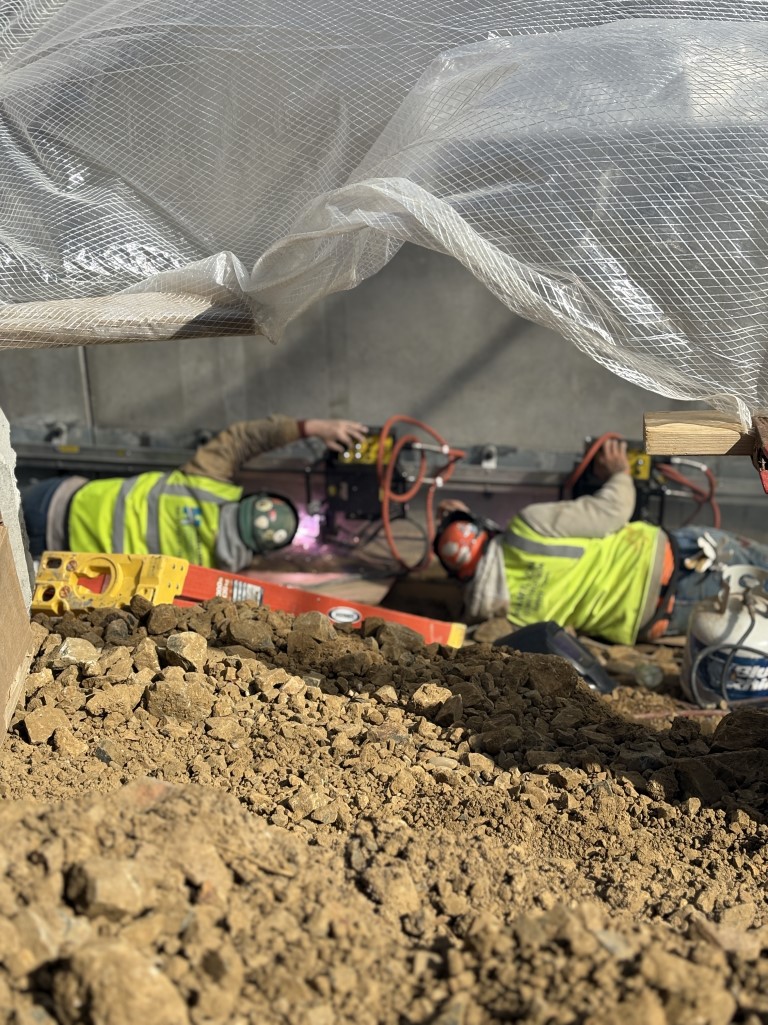 Two workers lying sideways welding at the base of a wall.