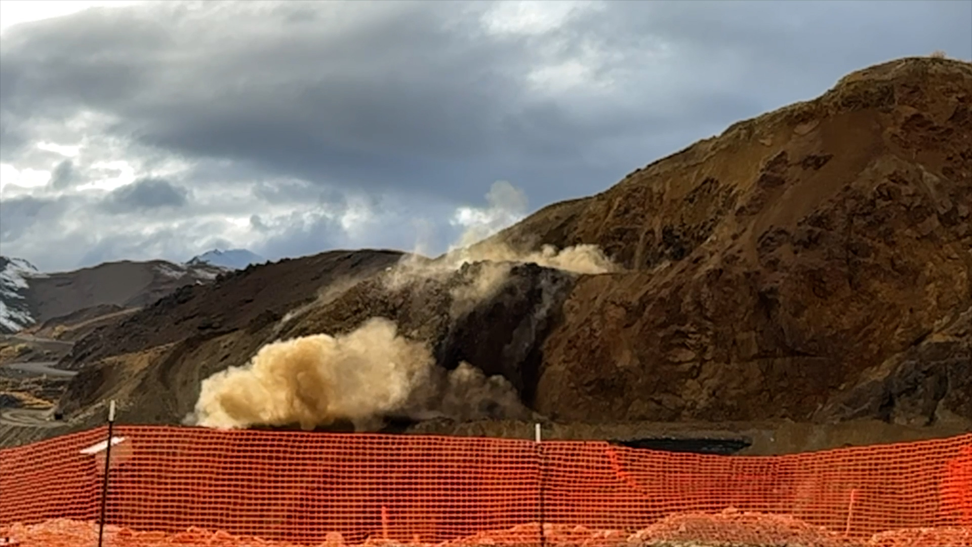 Behind an orange construction fence, a dust cloud rises from a rocky slope that has just been blasted.