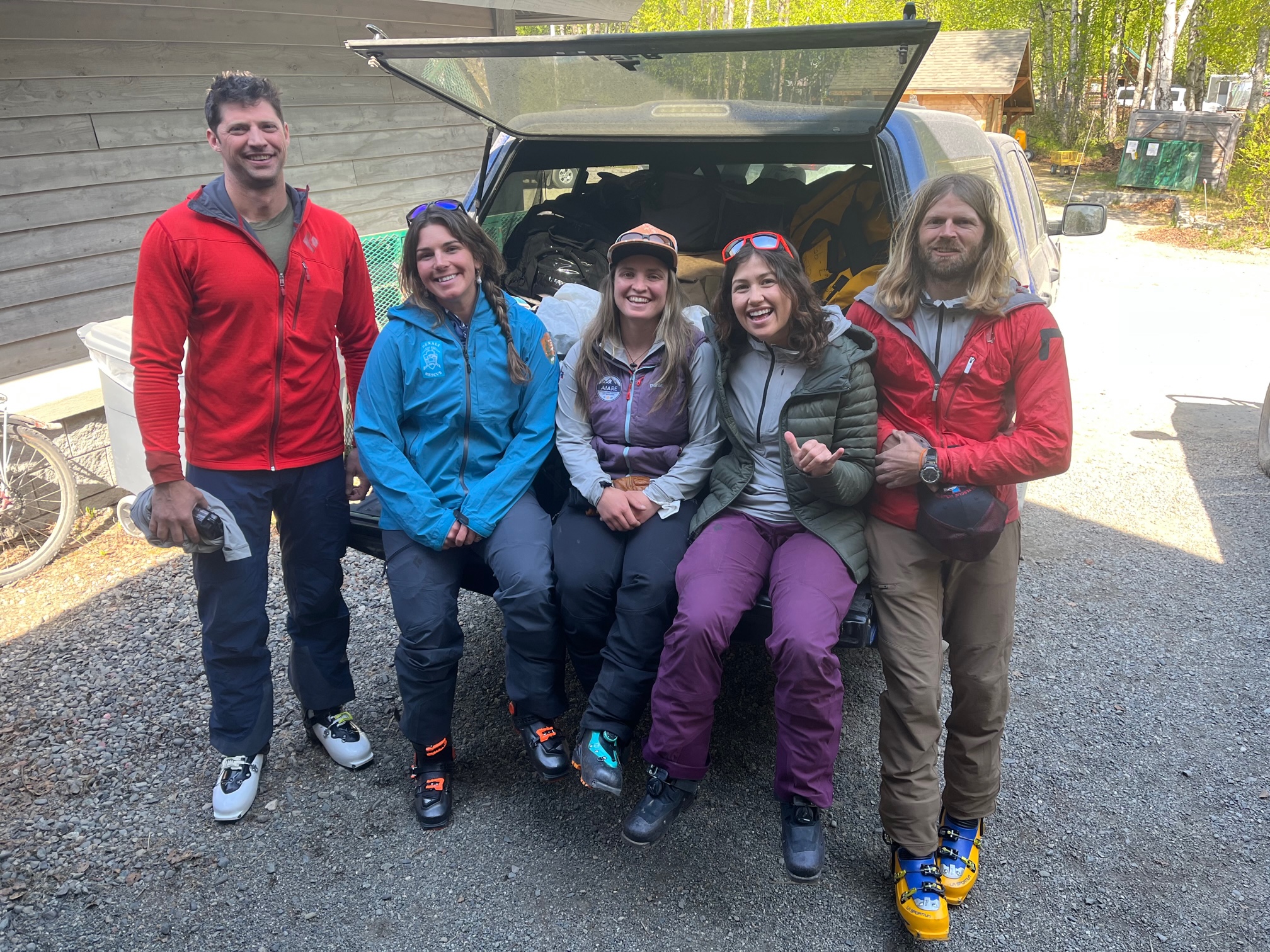 Five smiling climbers sit on the back of a pickup truck filled with duffel bags