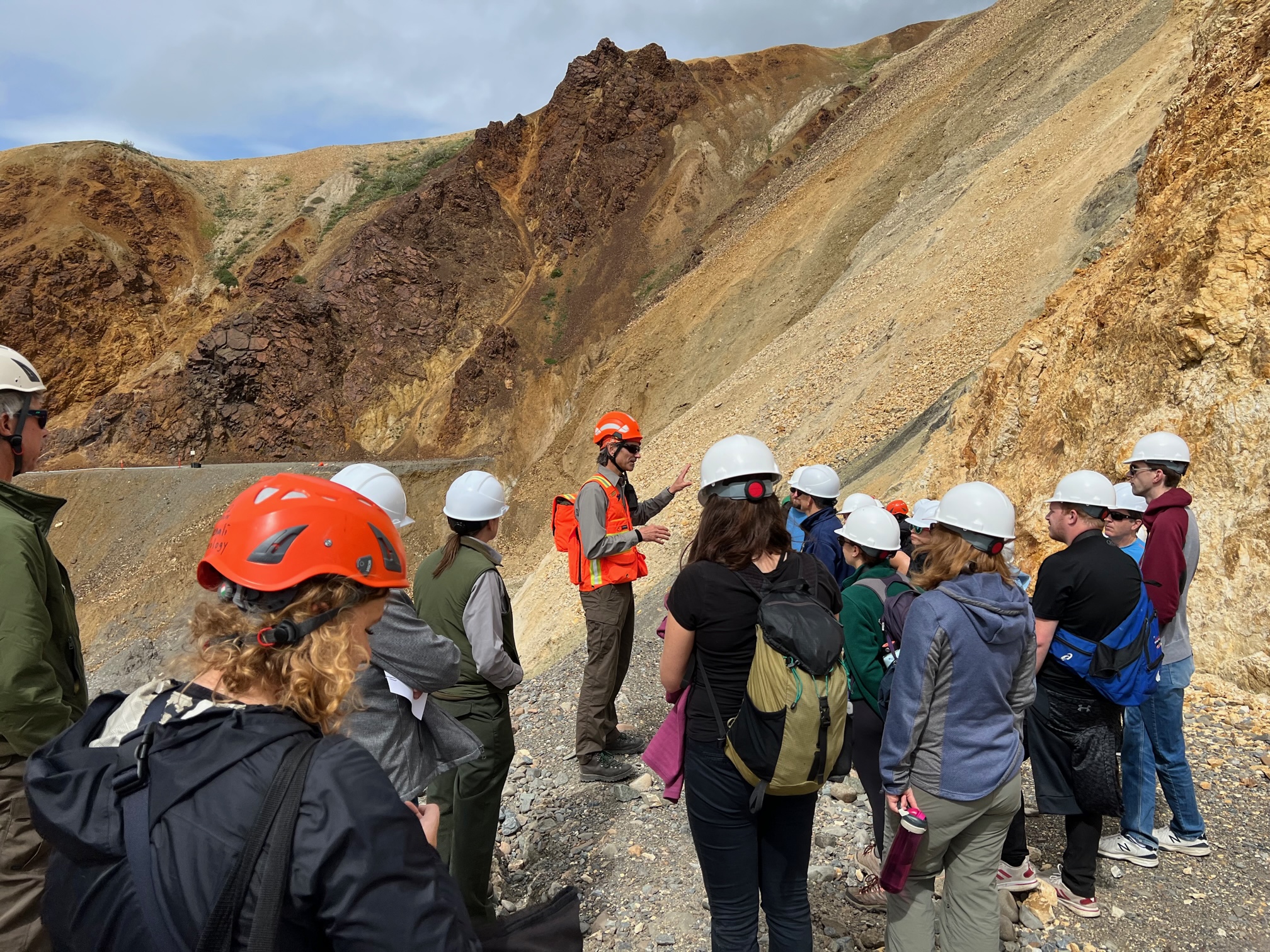 A group of about 15 people wearing protective hard hats stands at the edge of the Pretty Rocks landslide, where the gravel road bed drops away and slumps down a rocky mountainside. A few park staff are in uniform and one man speaks to the whole group.